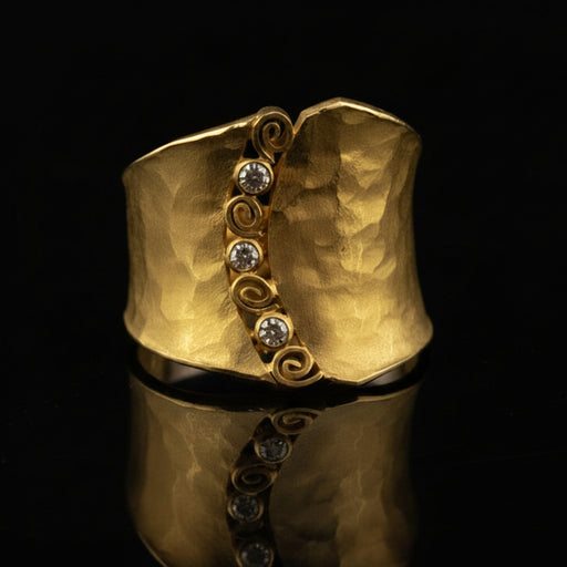 A hammered gold cuff ring with a vertical decorative band featuring four clear round-cut stones and swirl motifs, displayed on a reflective black surface.