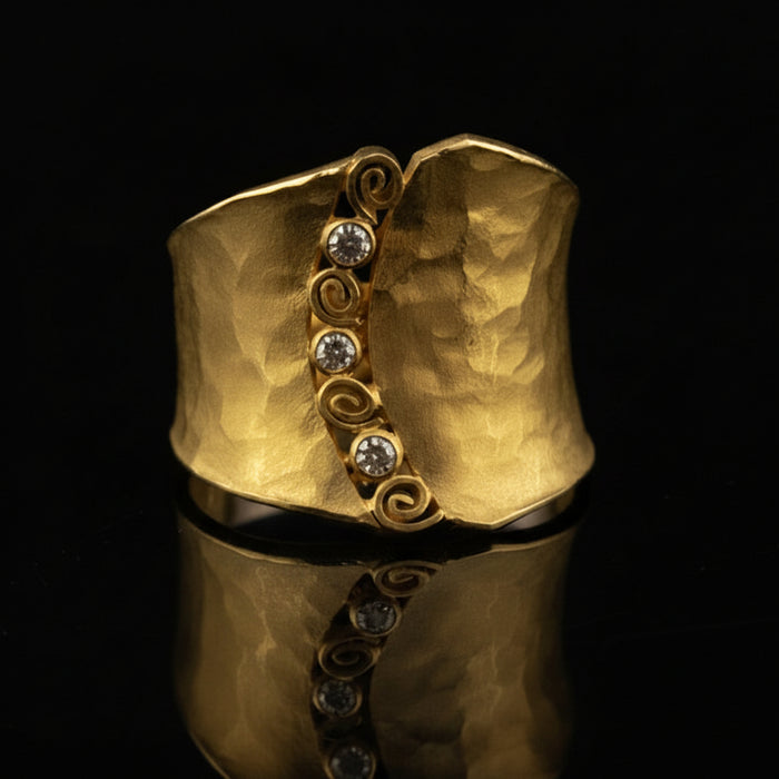 A hammered gold cuff ring with a vertical decorative band featuring four clear round-cut stones and swirl motifs, displayed on a reflective black surface.