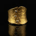 A hammered gold cuff ring with a vertical decorative band featuring four clear round-cut stones and swirl motifs, displayed on a reflective black surface.